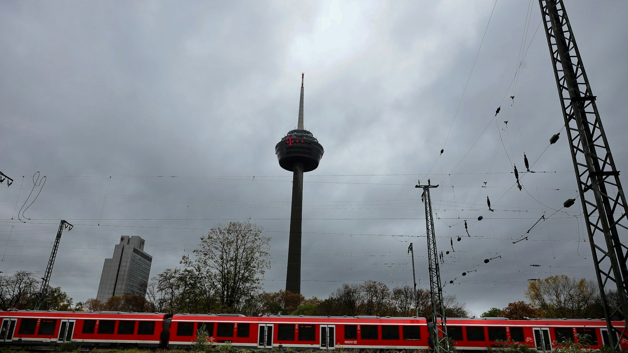 Der Kölner Fernsehturm Colonius ist von dunklen Unwetterwolken umgeben. Im Vordergrund fährt ein Regionalzug auf den Gleisen vorbei. (Archivbild)
