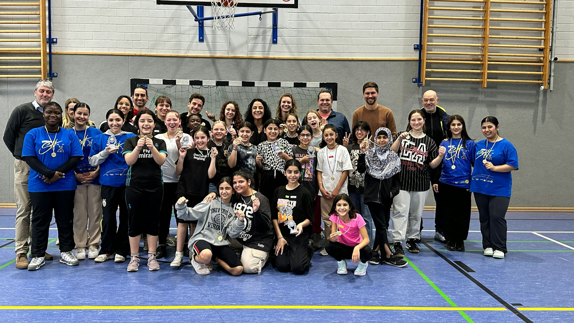 In einer Turnhalle posieren Schülerinnen mit Lehrer mit einer jungen Frau.