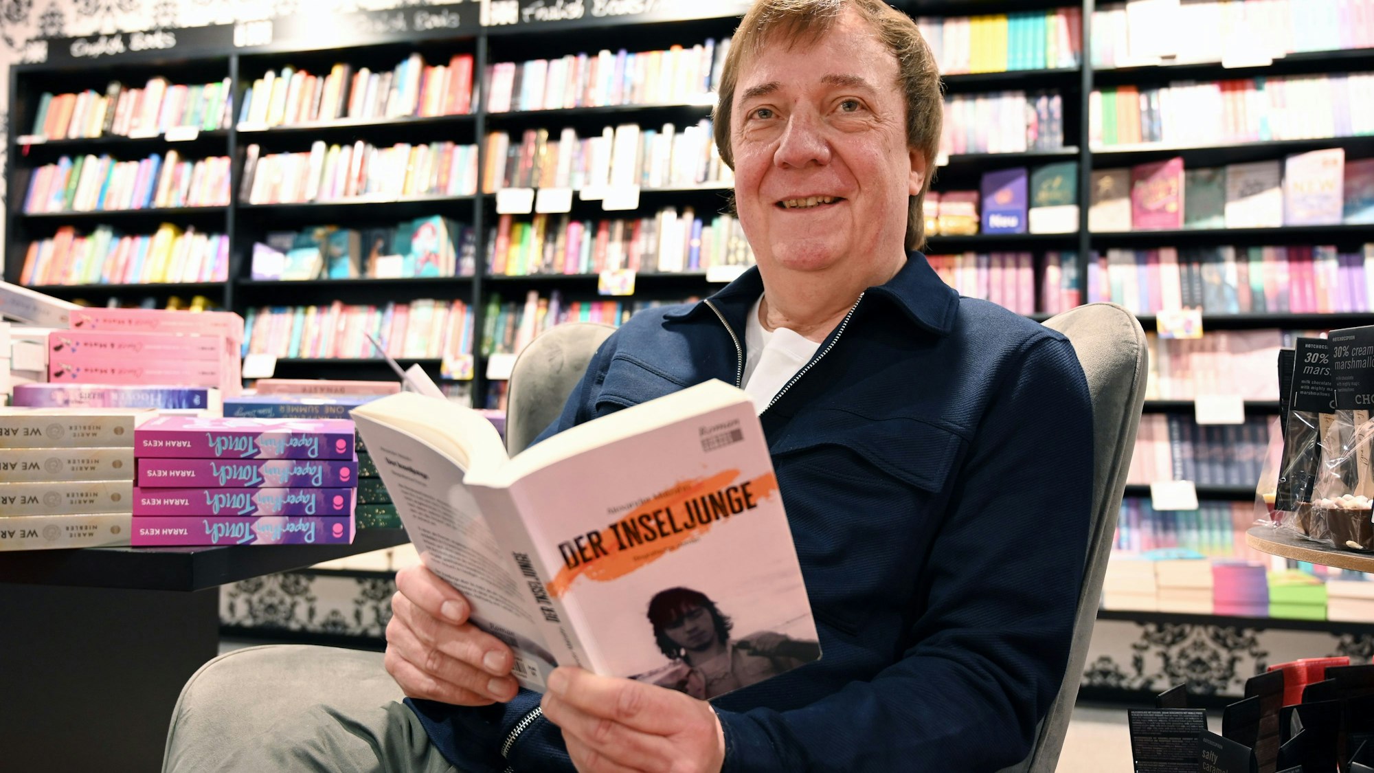 Ein Mann sitzt in einer Buchhandlung in einem Sessel. In der Hand hält er seinen eigenen Roman mit dem Titel „Der Inseljunge“.