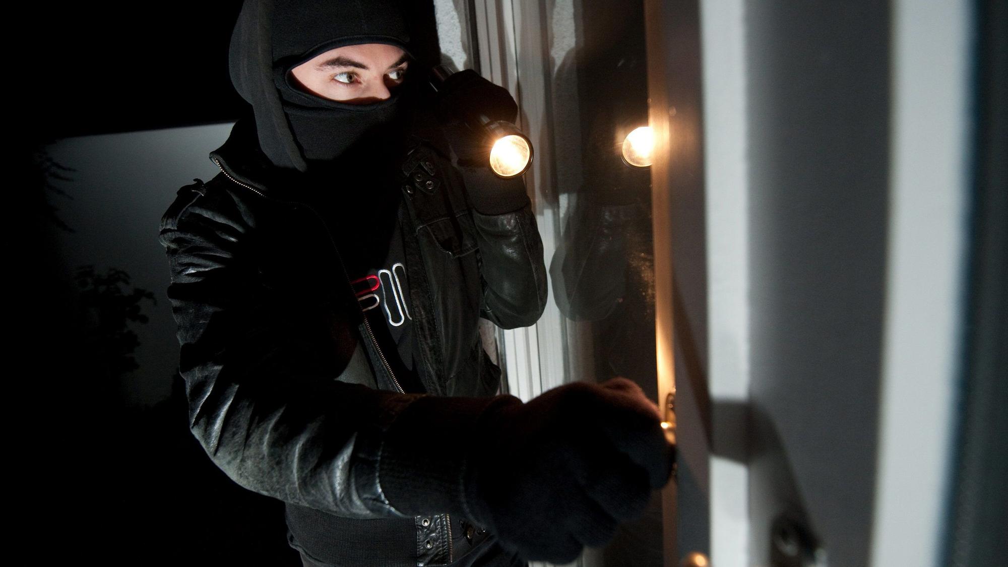 Ein vermummter Einbrecher mit Taschenlampe schaut durch ein Fenster in eine Wohnung.