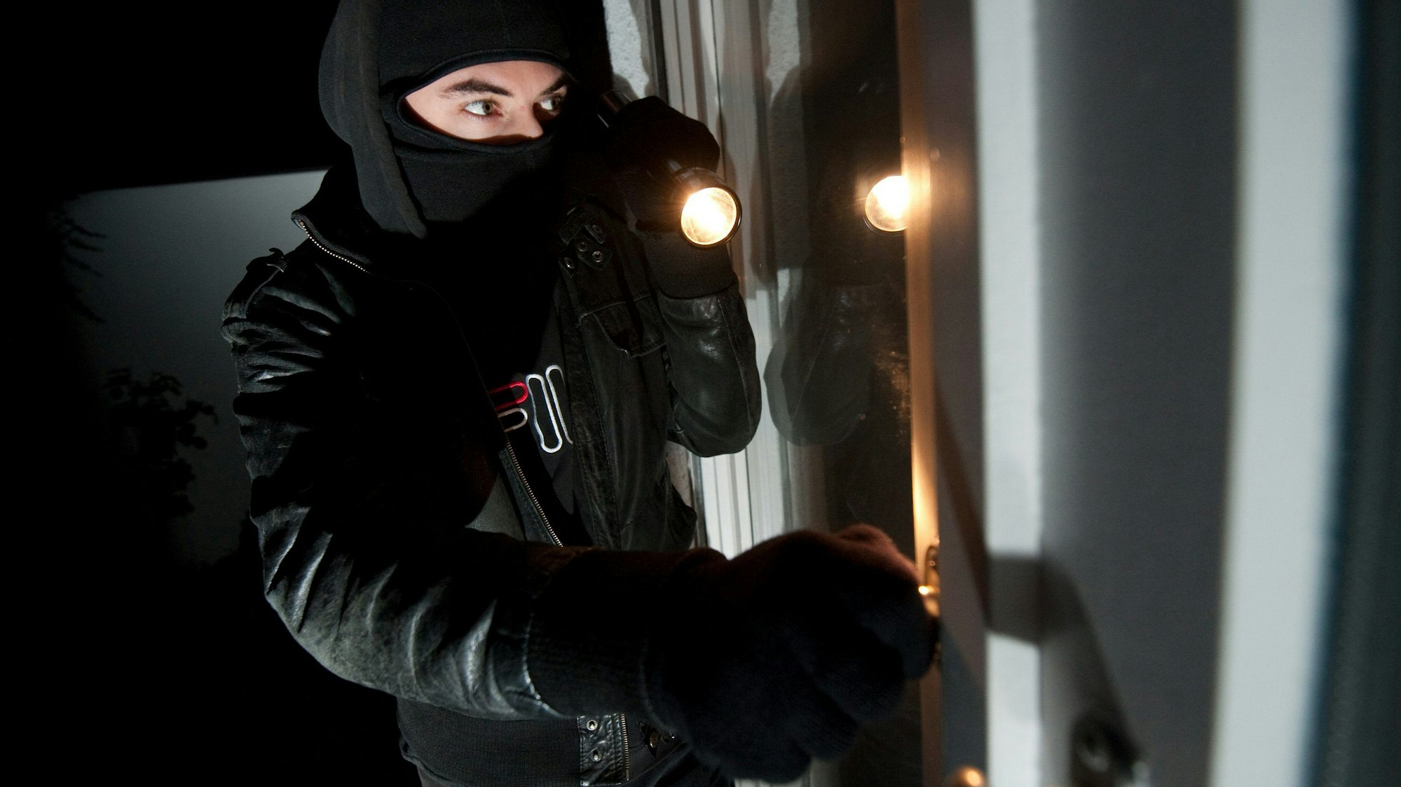 Ein vermummter Einbrecher mit Taschenlampe schaut durch ein Fenster in eine Wohnung.