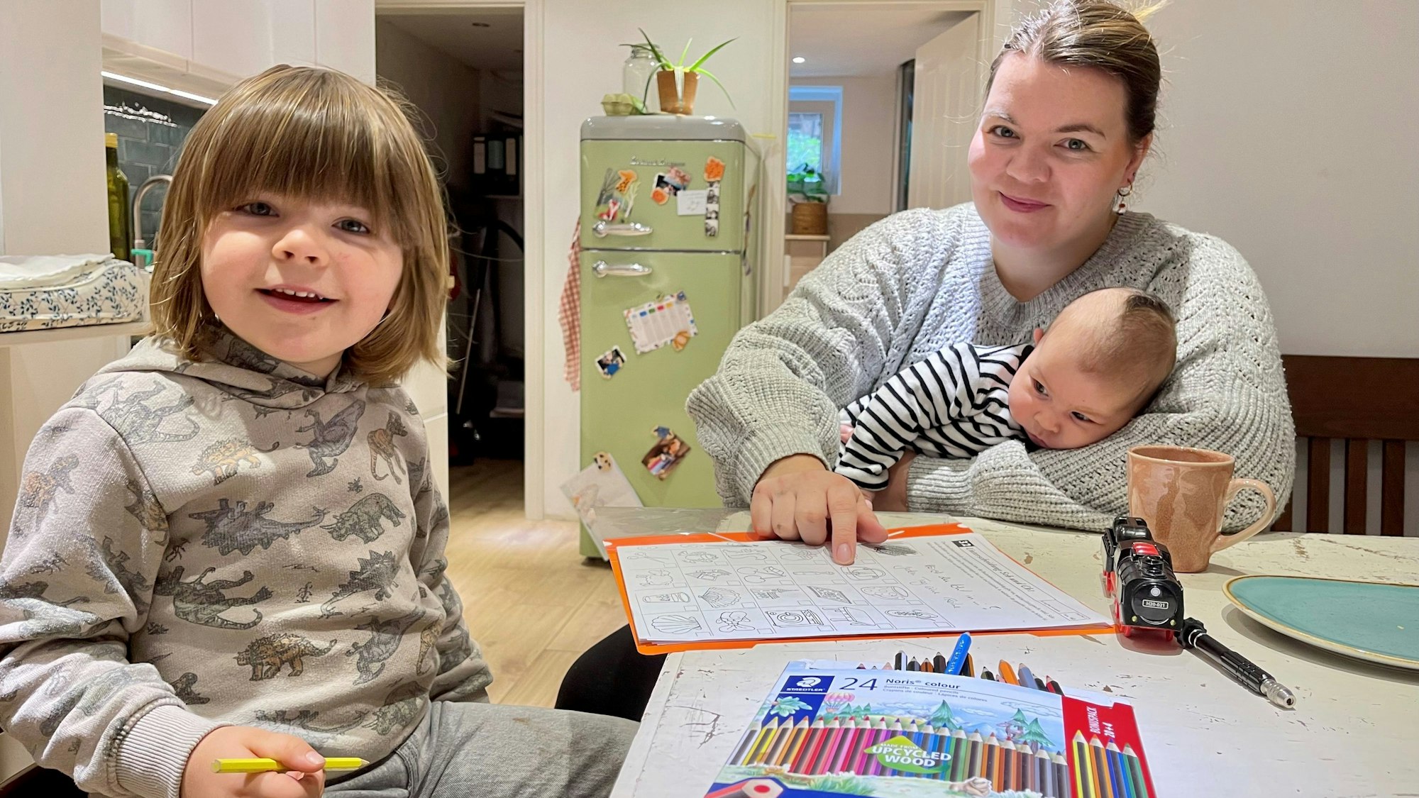 Eine Mutter sitzt mit ihren beiden Kindern an einem Tisch. Vor ihnen liegen Malstifte und Papier.