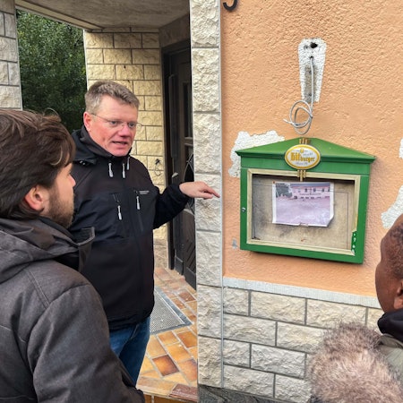 Das Bild zeigt Karl Kreuzberg vor der Gaststätte in Schweinheim. Menschen aus Ghana hören ihm zu.