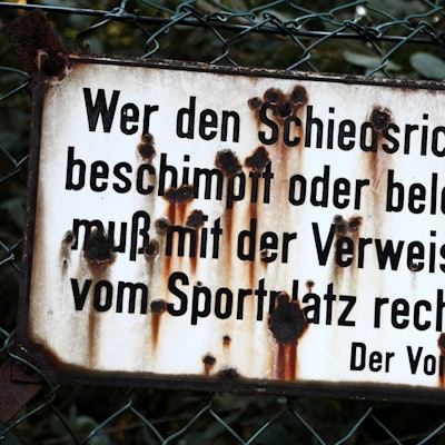 „Wer den Schiedsrichter beschimpft oder beleidigt, muß mit der Verweisung vom Sportplatz rechnen. Der Vorstand“ steht auf einem Schild an einem Sportplatz, das von Rost zerfressen ist.