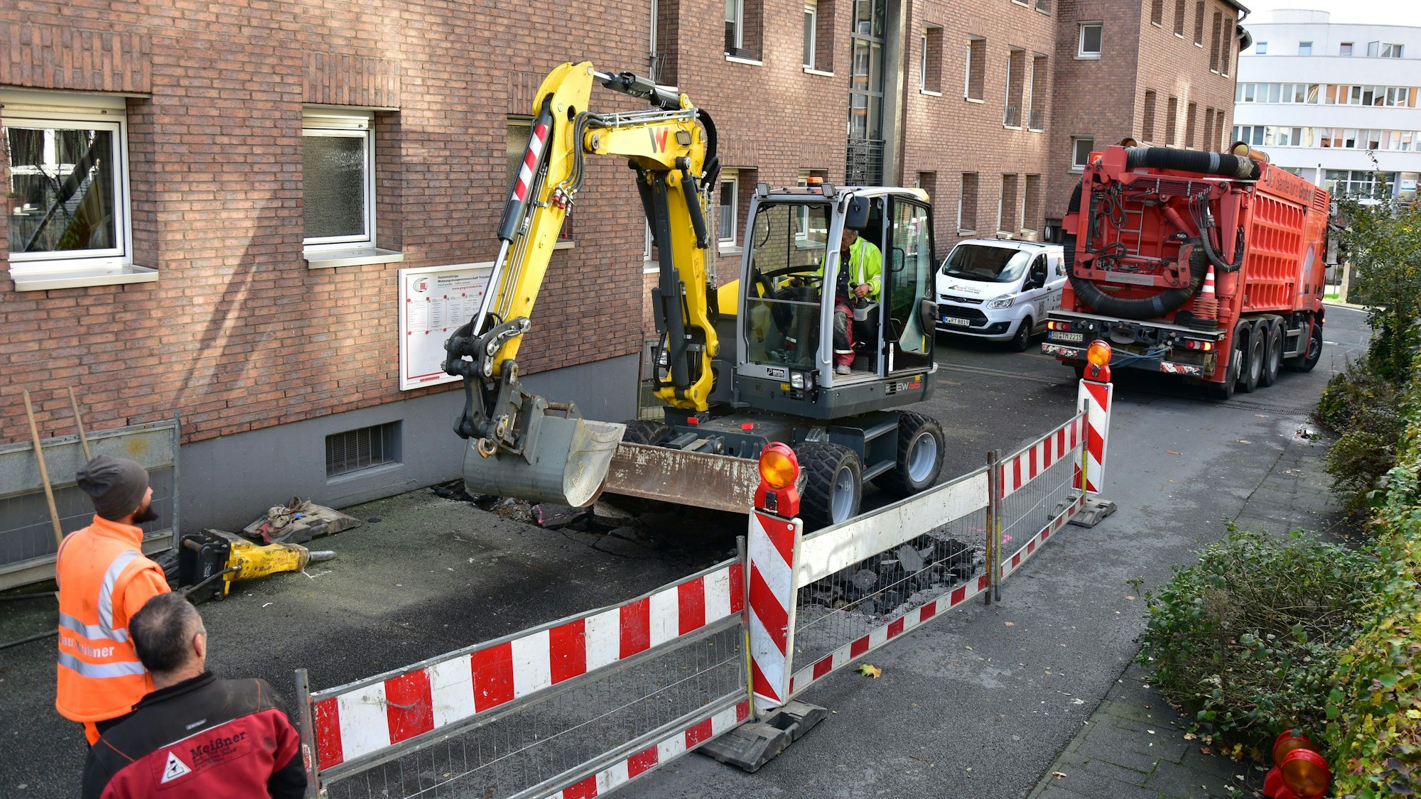 Baufahrzeuge auf einer Straße. Hinter rot-weißen Absperrungen arbeitet ein Bagger. Im Hintergrund steht ein rotes Tiefbaufahrzeug.