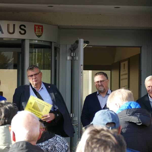 Vor dem Eingang zum Rathaus steht ein Mann, der eine gelbe Seite in der Hand hält. Vor ihm stehen mehrere Menschen, neben ihm eine ernste Frau mit kurzen Haaren.