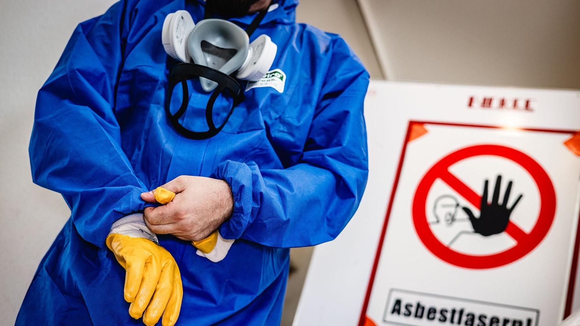 EIne Person steht vor einem Asbest-Warnschild und zieht sich Schutzhandschuhe an.