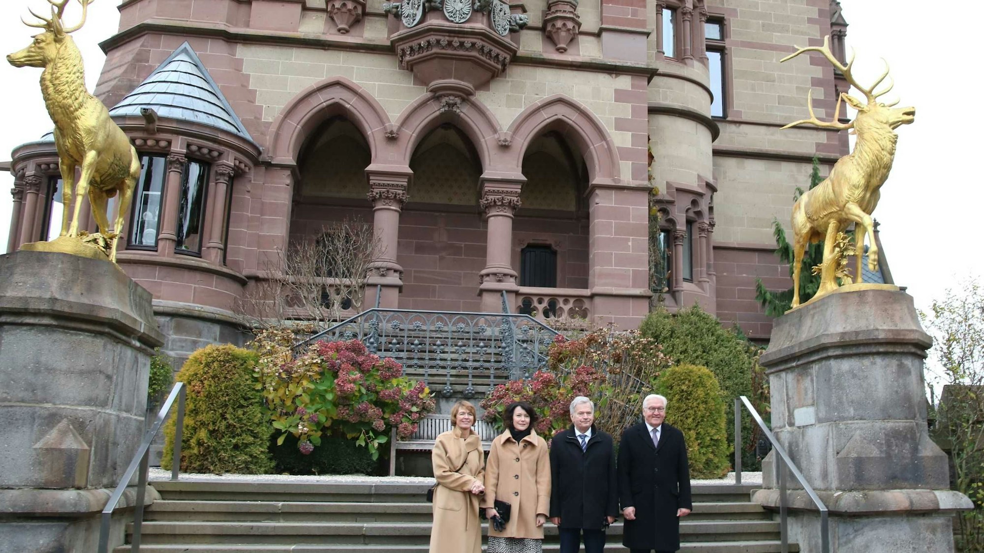 Die vier Personen stehen auf der Venusterrasse von Schloss Drachenburg, das historische Gemäuer im Hintergrund und flankiert von den zwei goldenen Hirschfiguren.