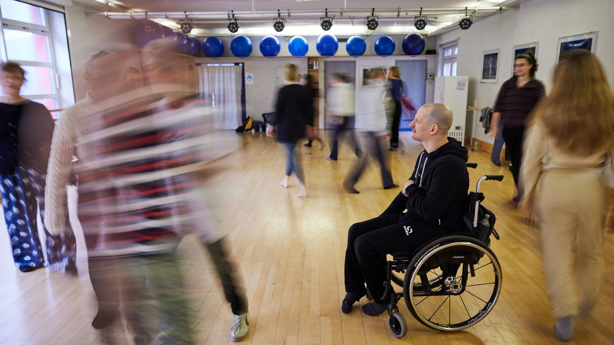 In einem Raum laufen Menschen scheinbar ziellos durcheinander, auch ein Mann im Rollstuhl ist dabei.