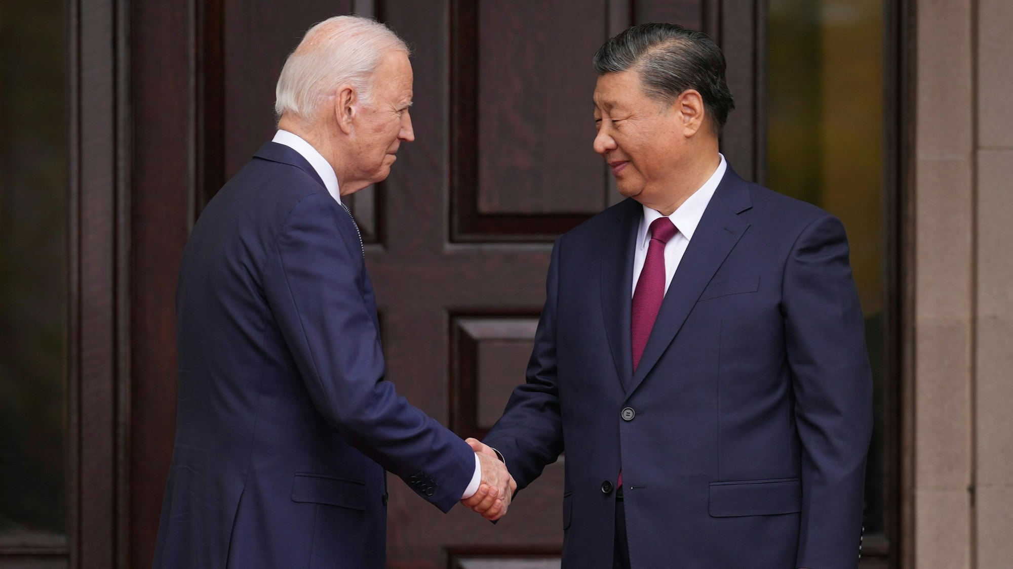 Joe Biden schüttelt die Hand von Xi Jinping, während die beiden sich im Anzug in die Augen schauen und vor einer hölzernen Tür stehen.