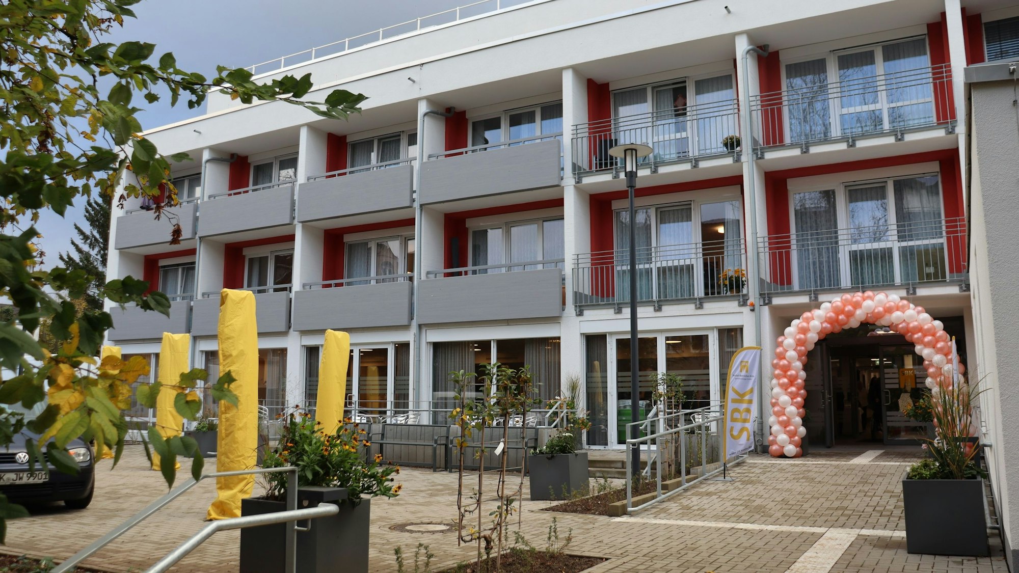 Ein dreistöckiger Flachbau mit einem von Luftballons geschmückten Eingang ist zu sehen.