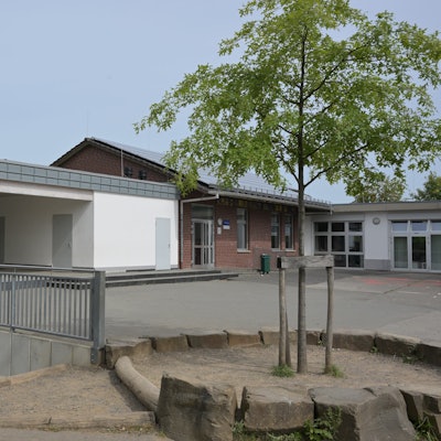 Der L-förmige Bau der KGS Burg Berge in Blecher formiert sich um den Pausenhof. Im Vordergrund ein Spielgelände mit einem jungen Laubbaum.