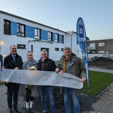 Eine Reihe von Menschen halten eine lange Pergamentpapierrolle vor einen zweigeschossigen Neubau.