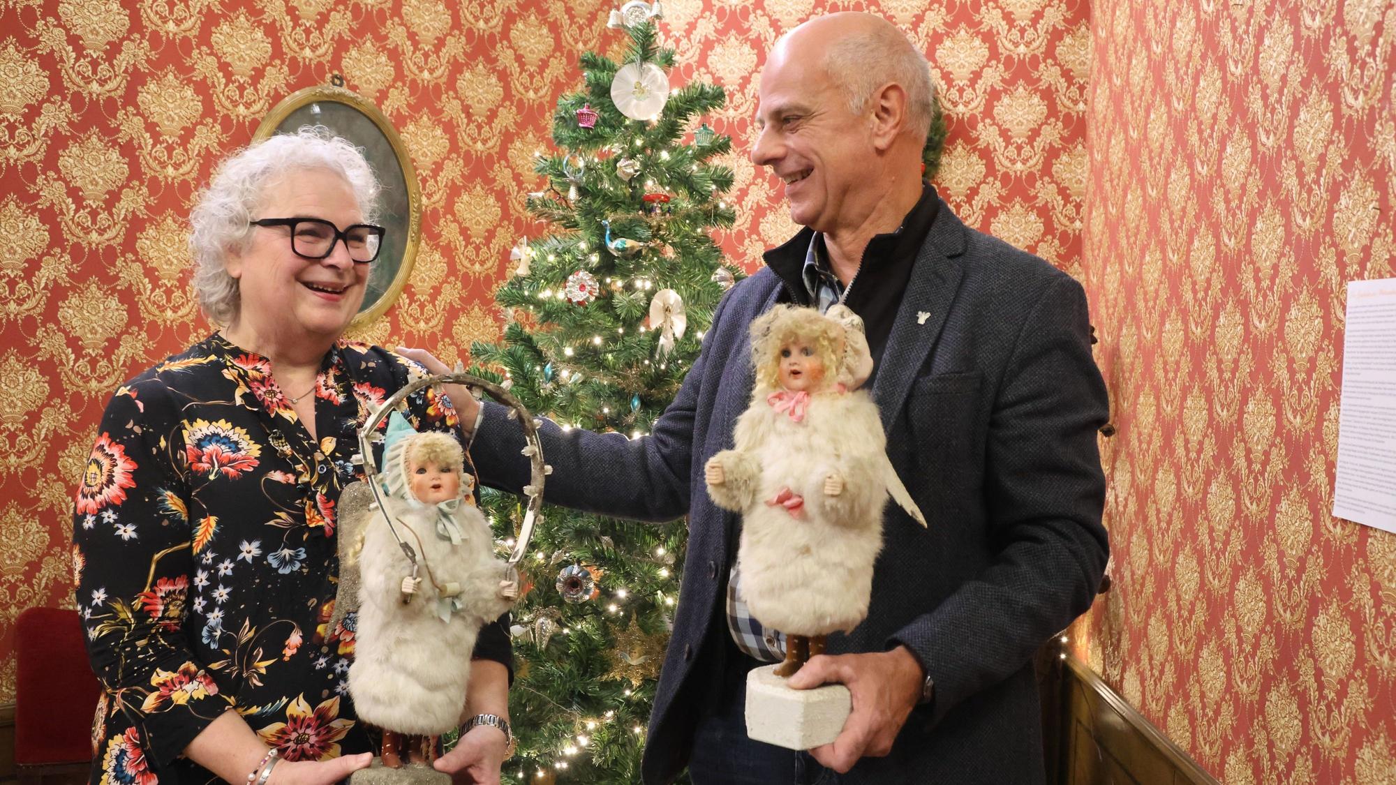 Eine Frau und ein Mann halten Engel-Figuren vor einem Weihnachtsbaum.