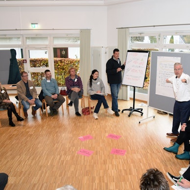 Unter der Moderation von Rainer Schmidt (rechts) ging es beim Auftaktworkshop in Wiehl-Oberbantenberg zunächst um das Verständnis von „Inklusion“. Hier steht der Moderator an einem Flipchart, die Teilnehmenden sitzen in einem Kreis um ihn herum.