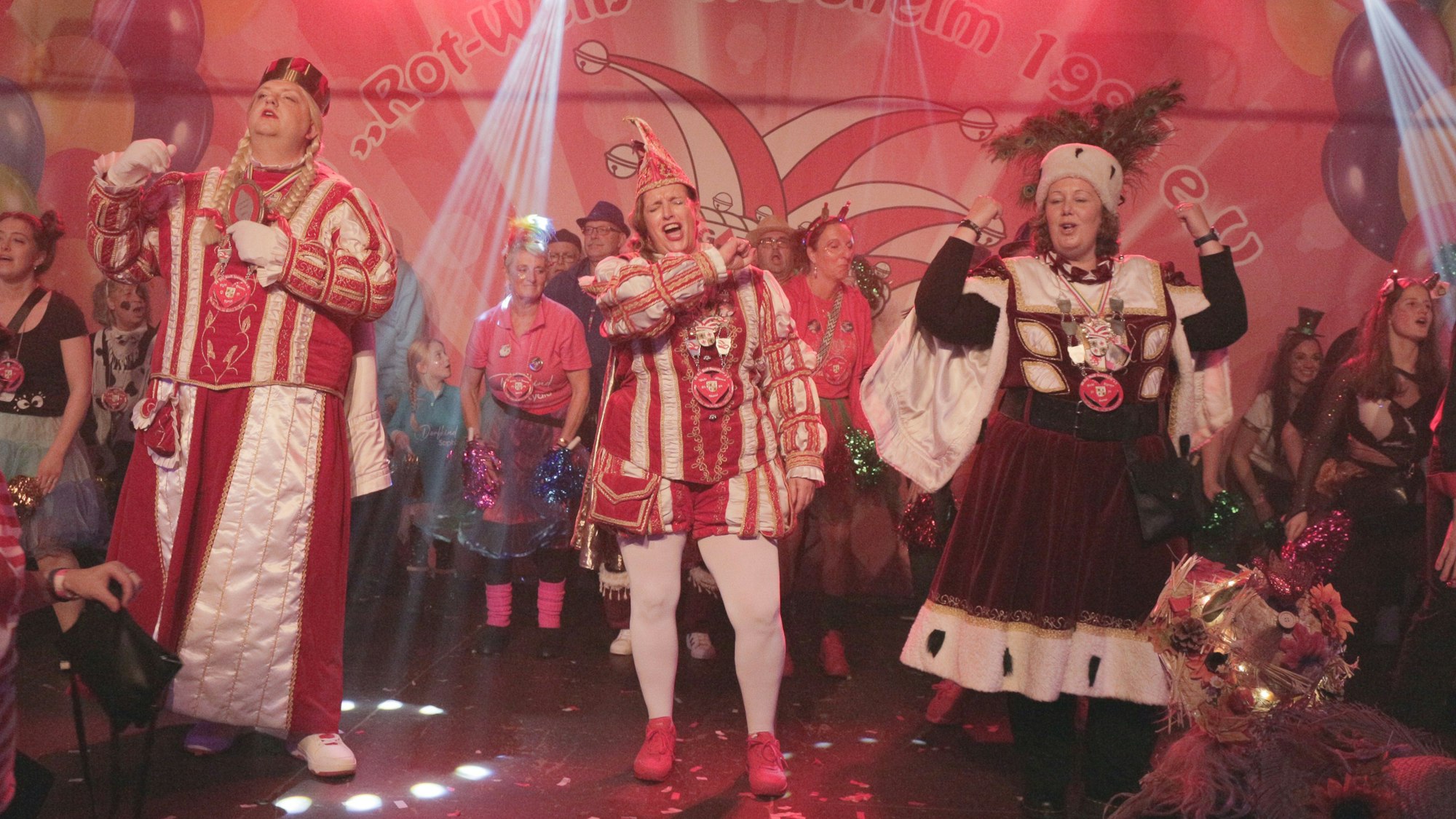 Die Mitglieder des Iversheimer Dreigestirns stehend singend auf der Bühne, hinter ihnen bunt kostümierte Karnevalsfreunde.