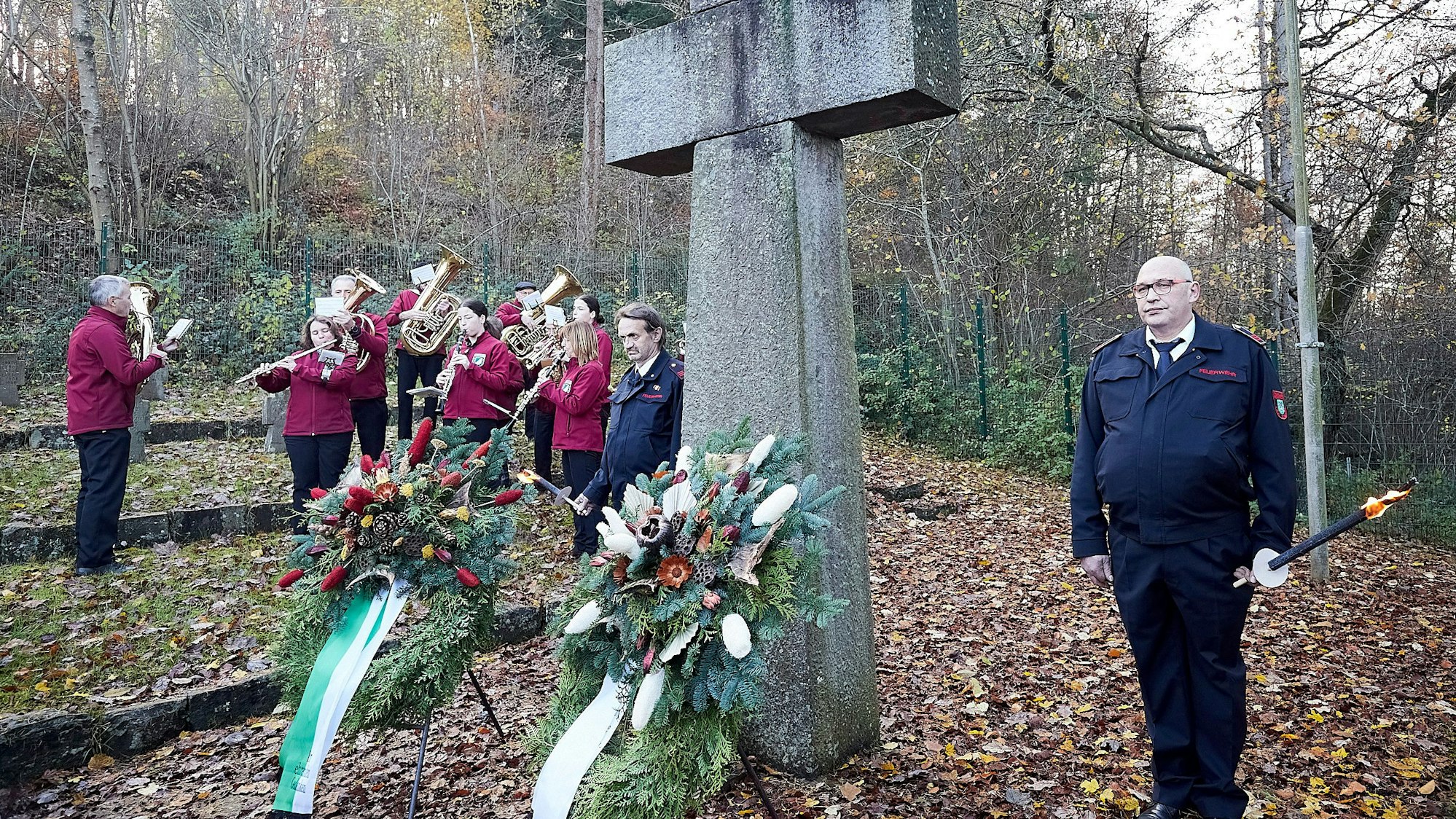 Eine Ehrenwache der Feuerwehr ist mit brennenden Pechfackeln am Kreuz des Ehrenfriedhofs in Schleiden angetreten. Im Hintergrund spielen die Ettelscheider Bergmusikanten.