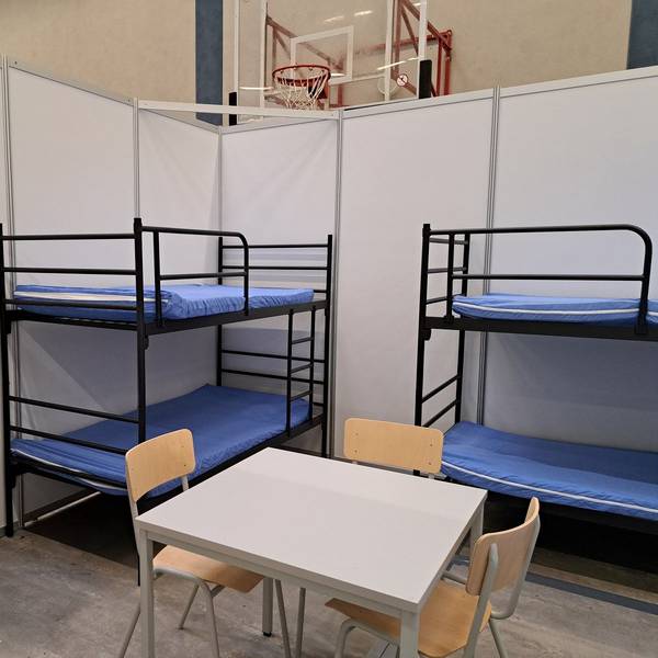 Zu sehen sind zwei Etagenbetten, ein Tisch, drei Stühle und ein grauer Schrank aus Metall.