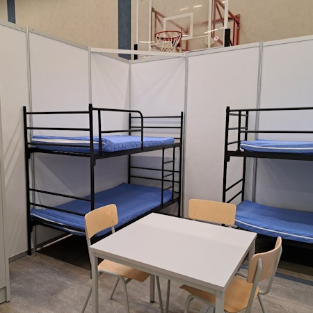 Zu sehen sind zwei Etagenbetten, ein Tisch, drei Stühle und ein grauer Schrank aus Metall.