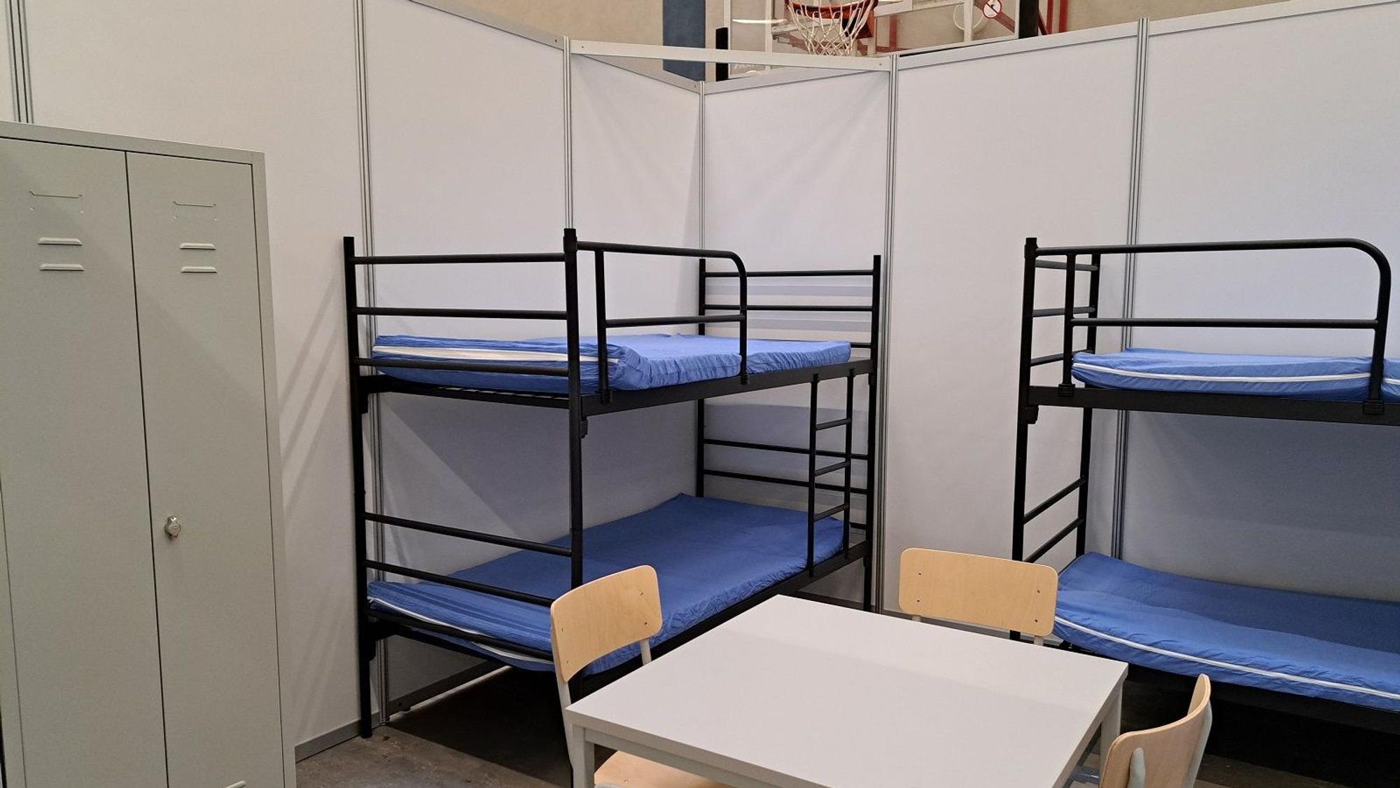 Zu sehen sind zwei Etagenbetten, ein Tisch, drei Stühle und ein grauer Schrank aus Metall.