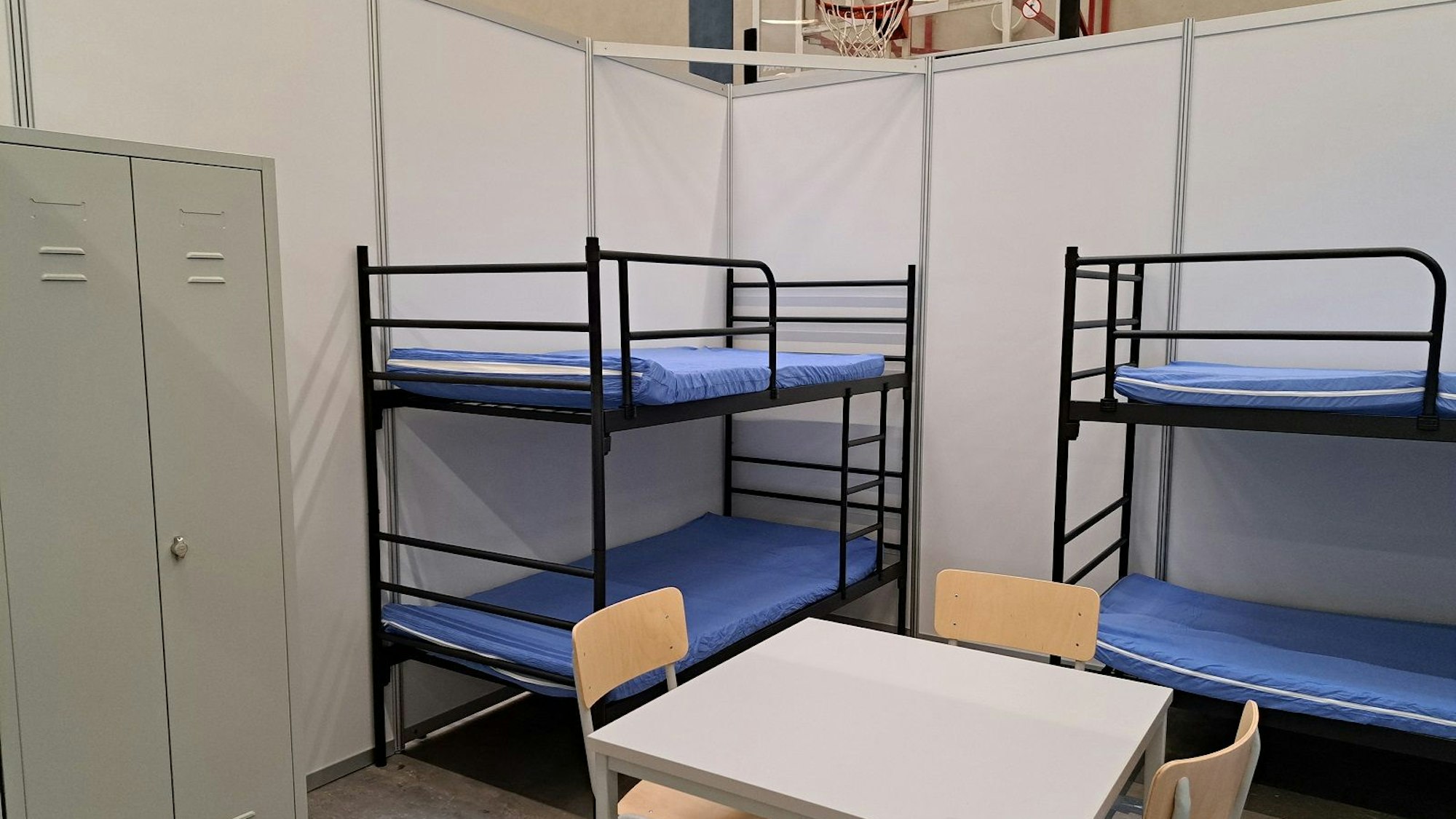 Zu sehen sind zwei Etagenbetten, ein Tisch, drei Stühle und ein grauer Schrank aus Metall.