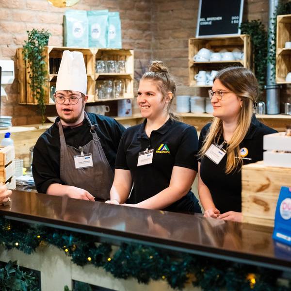 Drei junge Menschen hinter einem Tresen im Gespräch mit Ministerin Dorothee Feller.