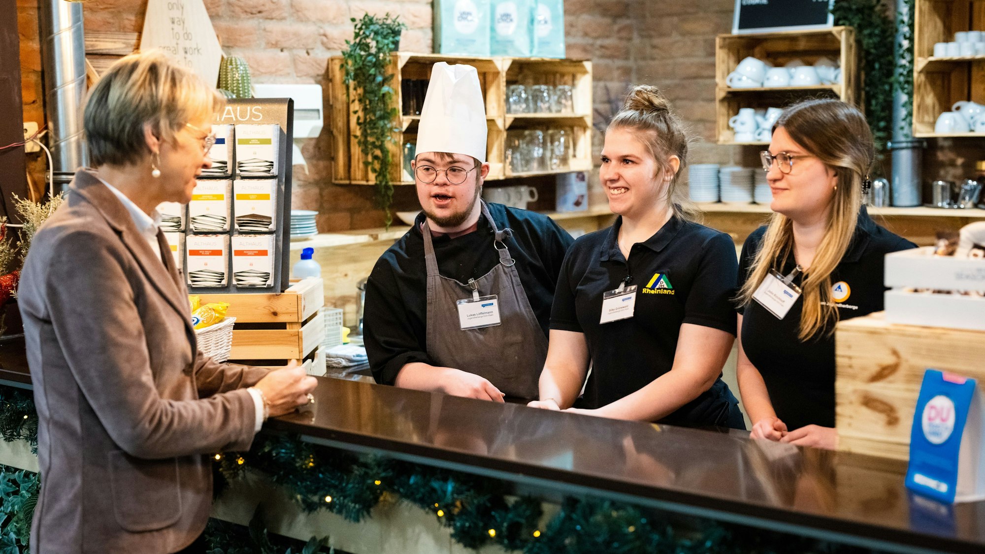 Drei junge Menschen hinter einem Tresen im Gespräch mit Ministerin Dorothee Feller.