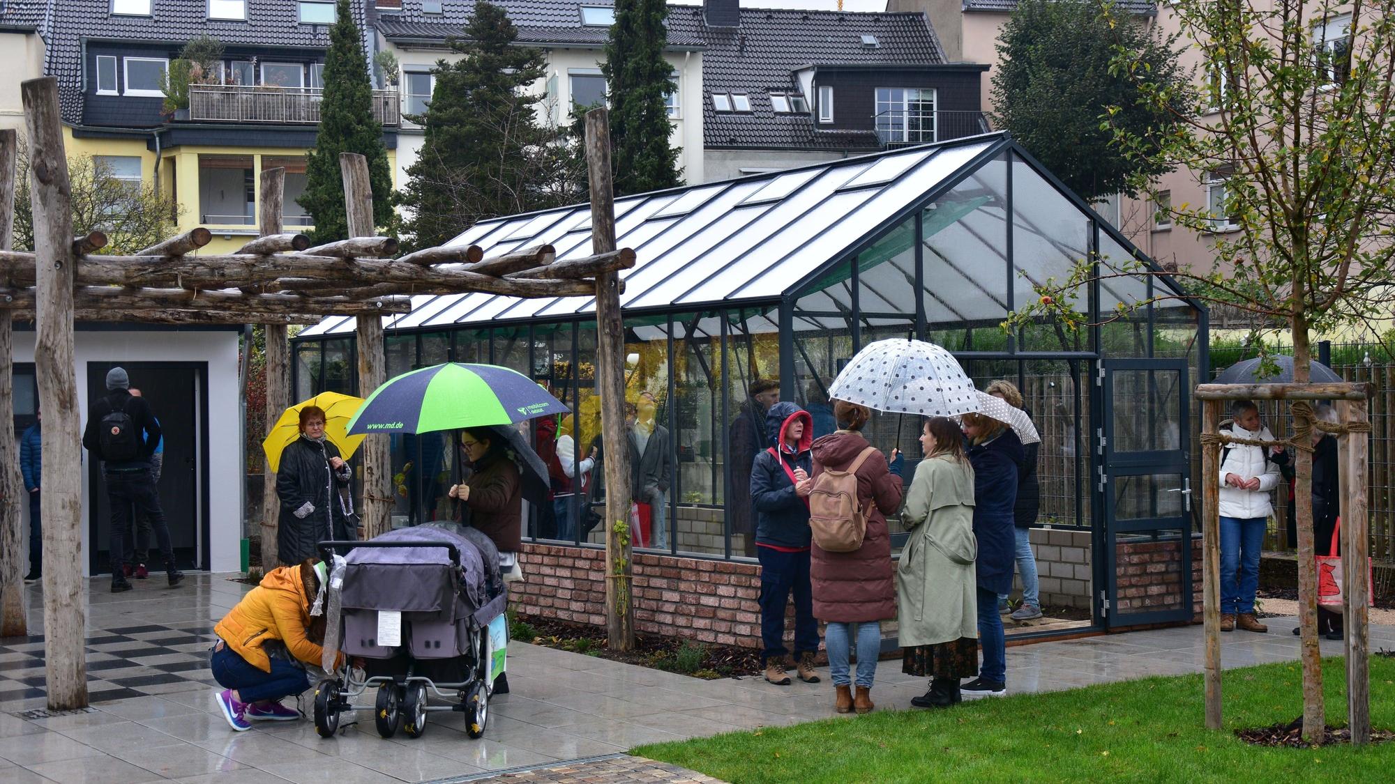 Leute mit Regenschirmen stehen vor Gewächshaus. Wohnhäuser im Hintergrund.