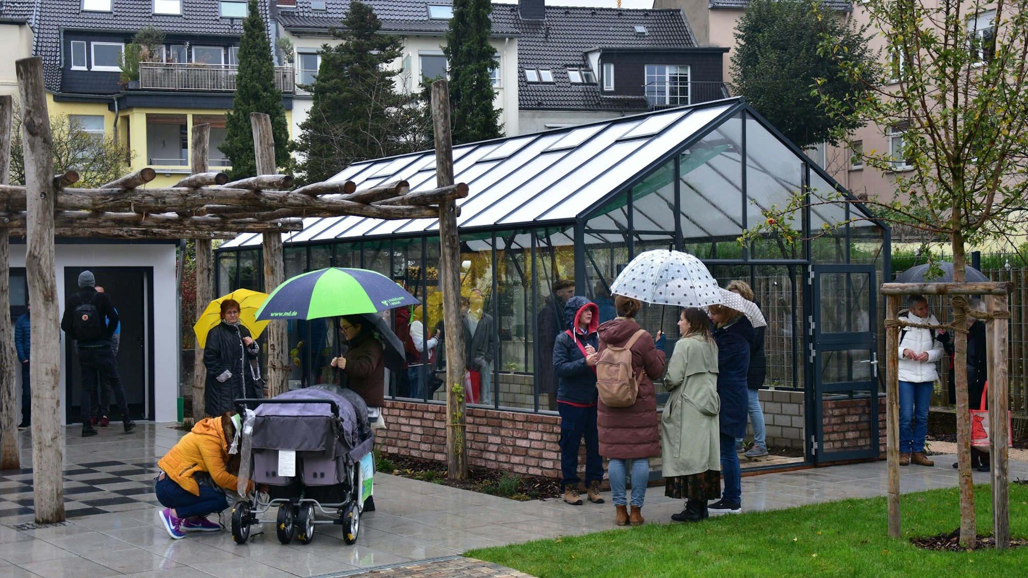 Leute mit Regenschirmen stehen vor Gewächshaus. Wohnhäuser im Hintergrund.