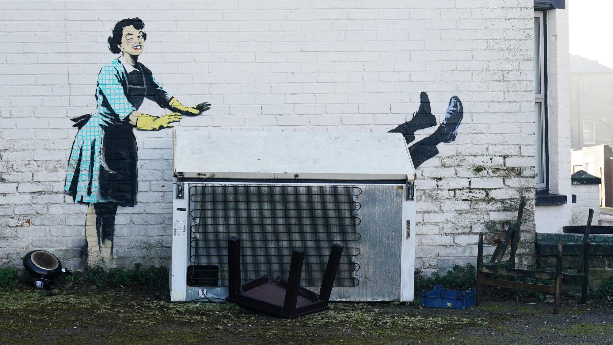 Blick auf das Kunstwerks „Valentine's Day Mascara“ des Künstlers Banksy an der Seite eines Gebäudes. Das Kunstwerk zeigt eine Hausfrau aus den 1950er Jahren, die eine klassische blaue Schürze und gelbe Spülhandschuhe trägt, ein geschwollenes Auge und einen fehlenden Zahn hat und ihren Partner scheinbar in eine Gefriertruhe schiebt.