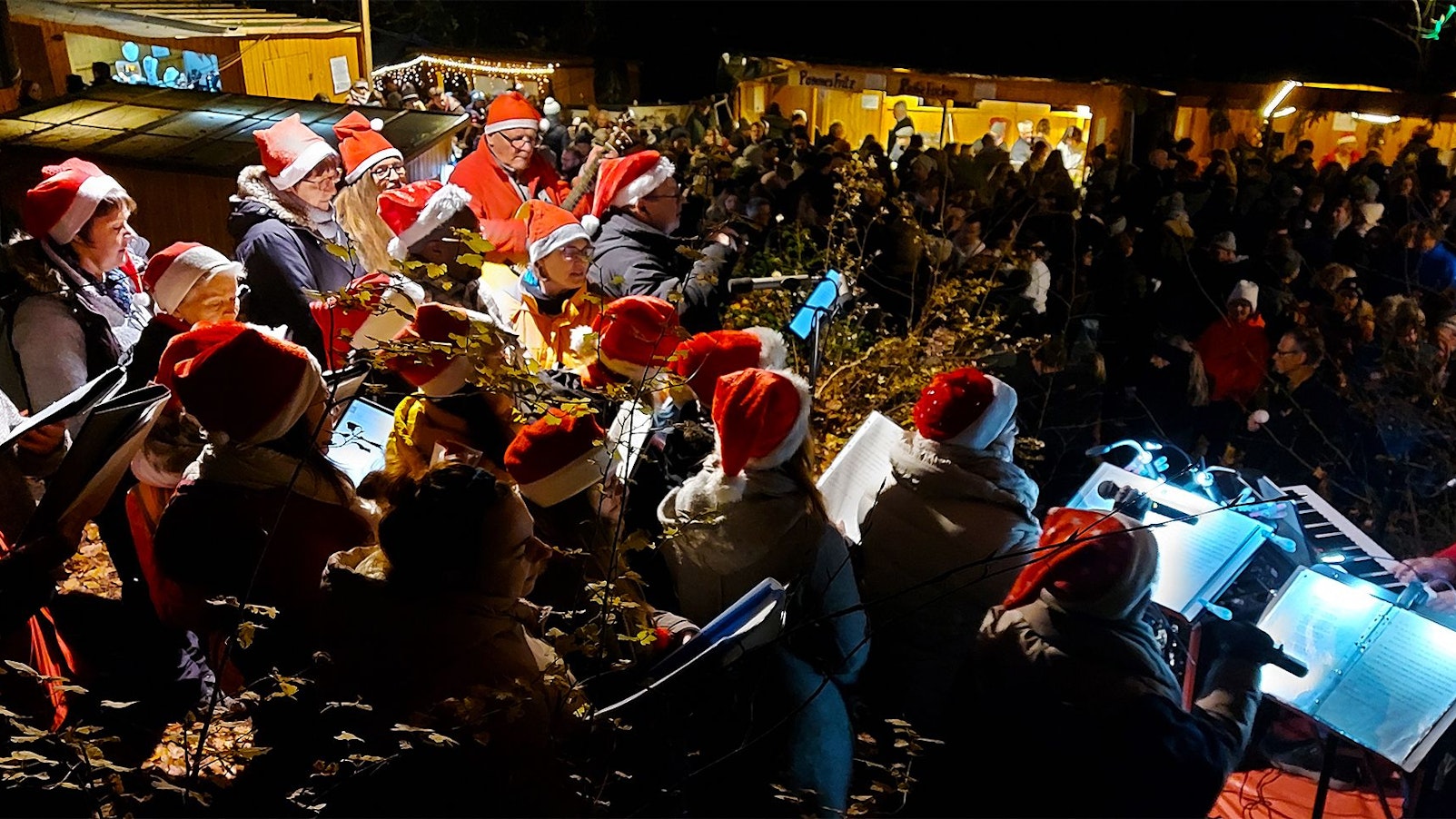 Menschen mit Nikolausmützen stehen im Dunkeln auf einem Weihnachtsmarkt und halten Noten in der Hand.