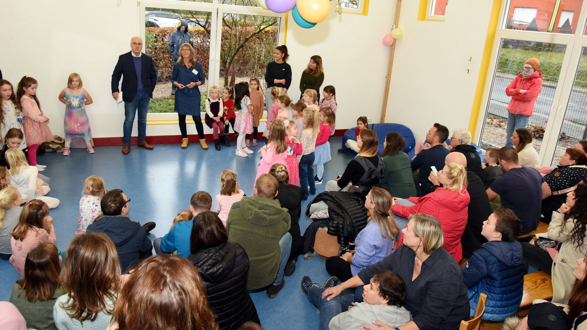 In einem großen Raum sind viele Kinder und Erwachsene unter bunten Luftballons versammelt.