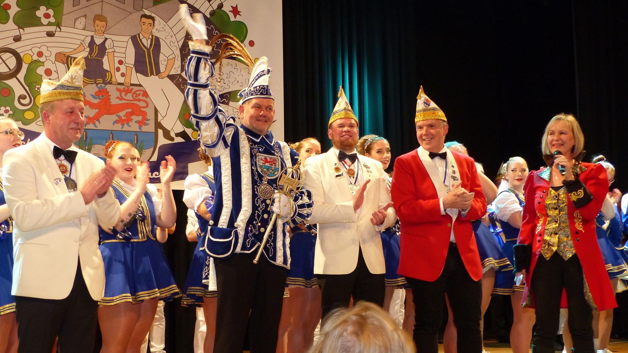 Auf einer Bühne stehen Mitglieder einer Karnevalsgesellschaft. In ihrer Mitte steht der Prinz und winkt ins Publikum.