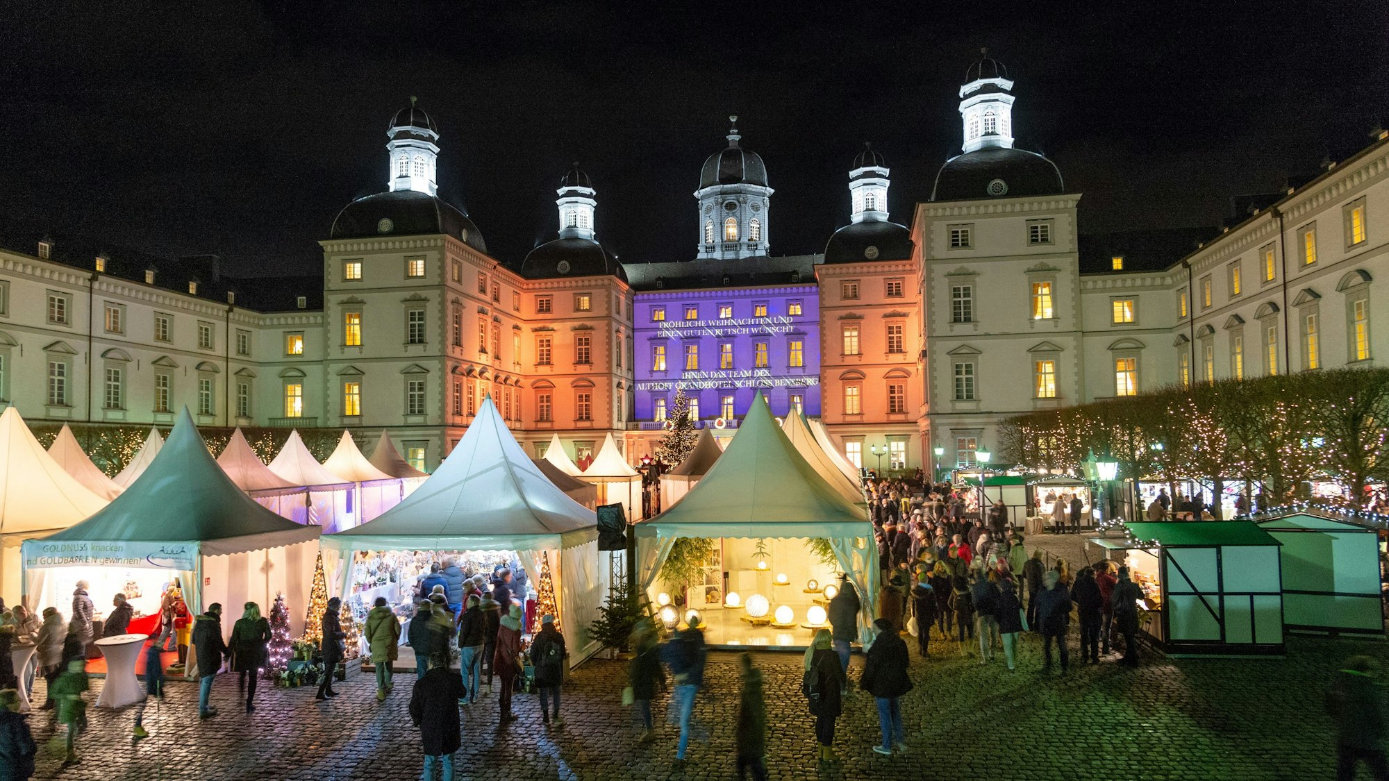 Bunt angeleuchtet werden die Mauern des Schloss Bensberg. Davor zahlreiche Weihnachtsmarktbuden.