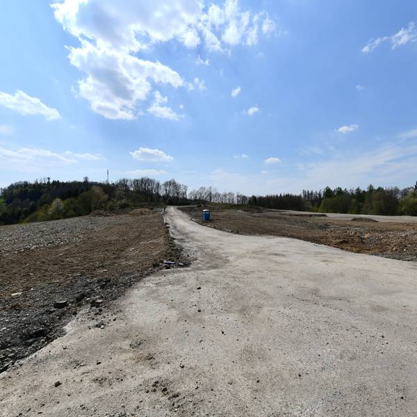 Ein leeres Feld aus Sand und Erde, in der Mitte ein Weg und ein Mülleimer. Im Hintergrund einige laublose Bäume vor strahlend blauem Himmel.