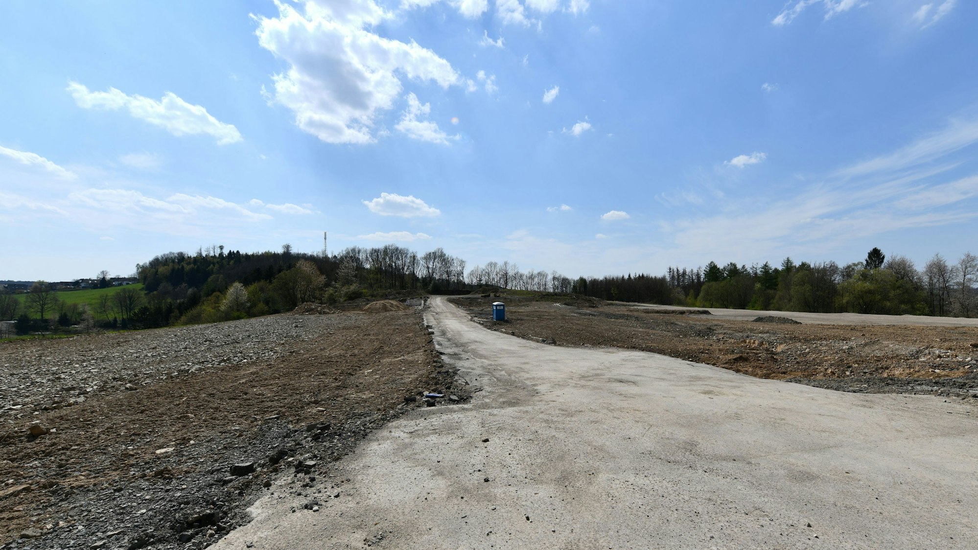 Ein leeres Feld aus Sand und Erde, in der Mitte ein Weg und ein Mülleimer. Im Hintergrund einige laublose Bäume vor strahlend blauem Himmel.