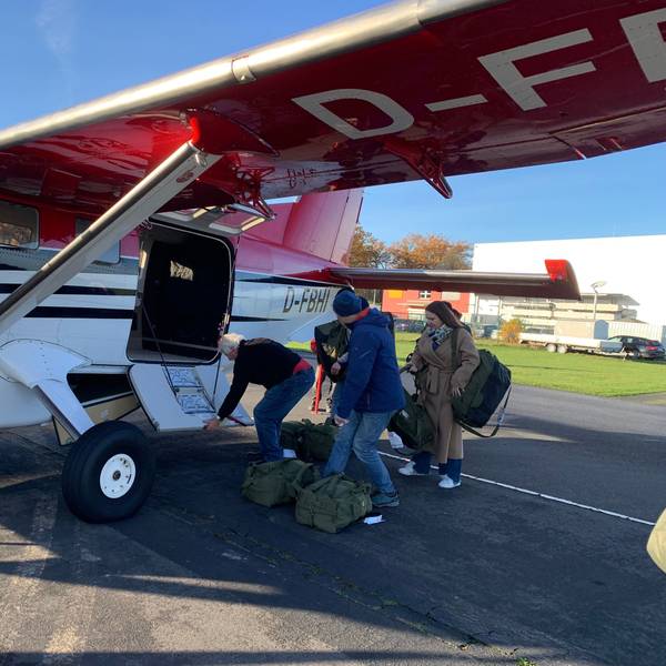 Drei Menschen stehen vor einem kleinen Flugzeug und beladen es mit gepackten Rucksäcken aus olivgrünem Stoff.
