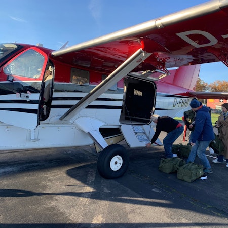 Drei Menschen stehen vor einem kleinen Flugzeug und beladen es mit gepackten Rucksäcken aus olivgrünem Stoff.