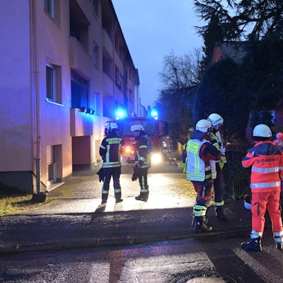 Feuerwehrleute stehen am Abend mit Blaulicht an ihren Einsatzfahrzeugen vor einem Mehrfamilienhaus.