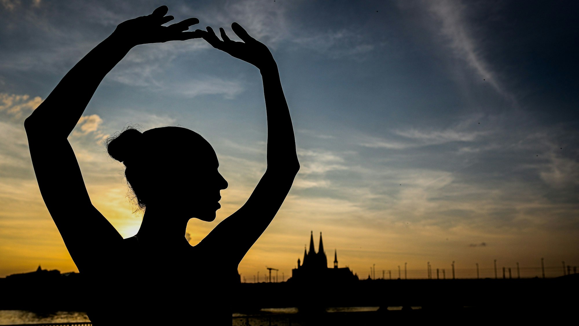 Ein Schatten einer Tänzerin vor blau-goldenen Himmel mit Sonnenuntergang, im Hintergrund die Silhouette des Doms.