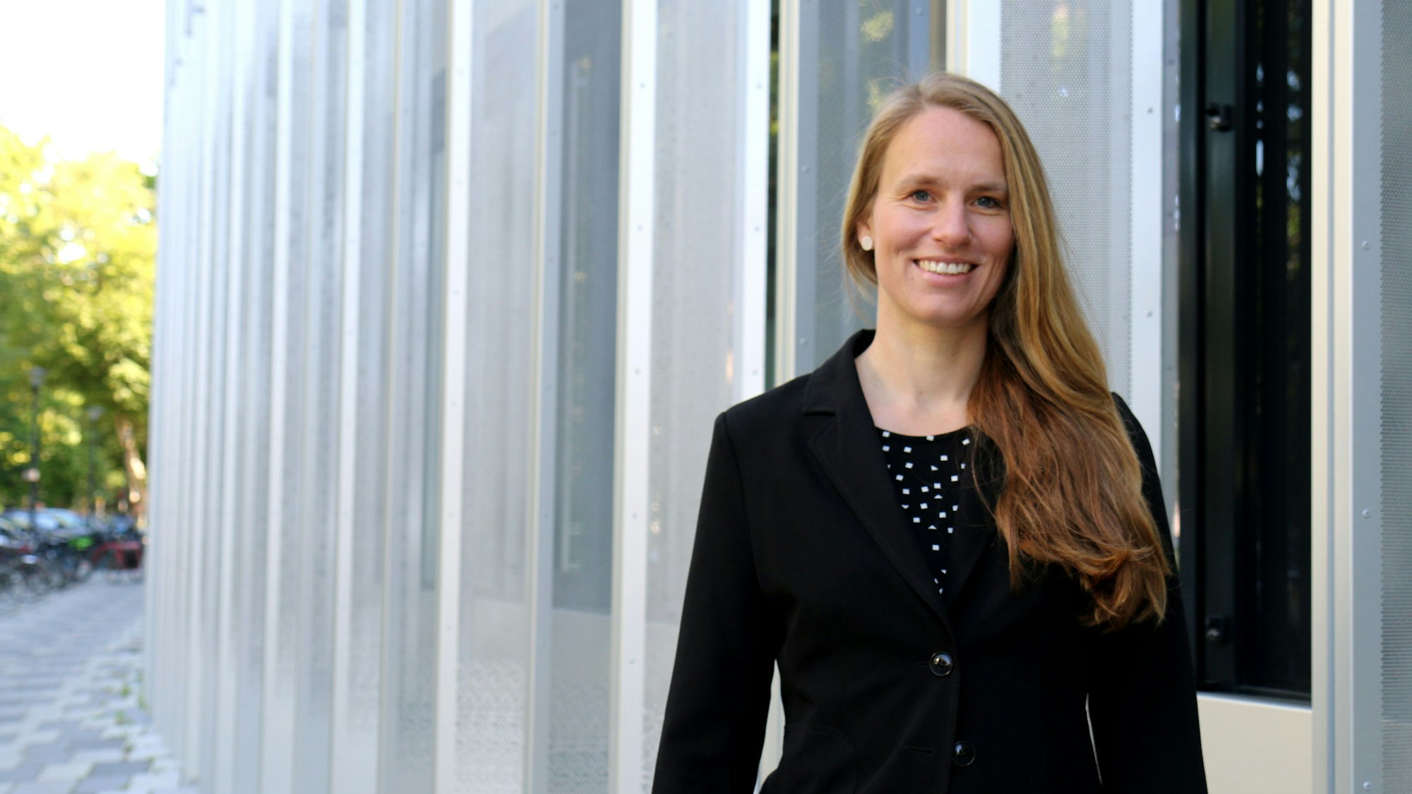 Die blonde Frau mit langen Haaren steht vor einem Gebäude mit moderner Fassade.