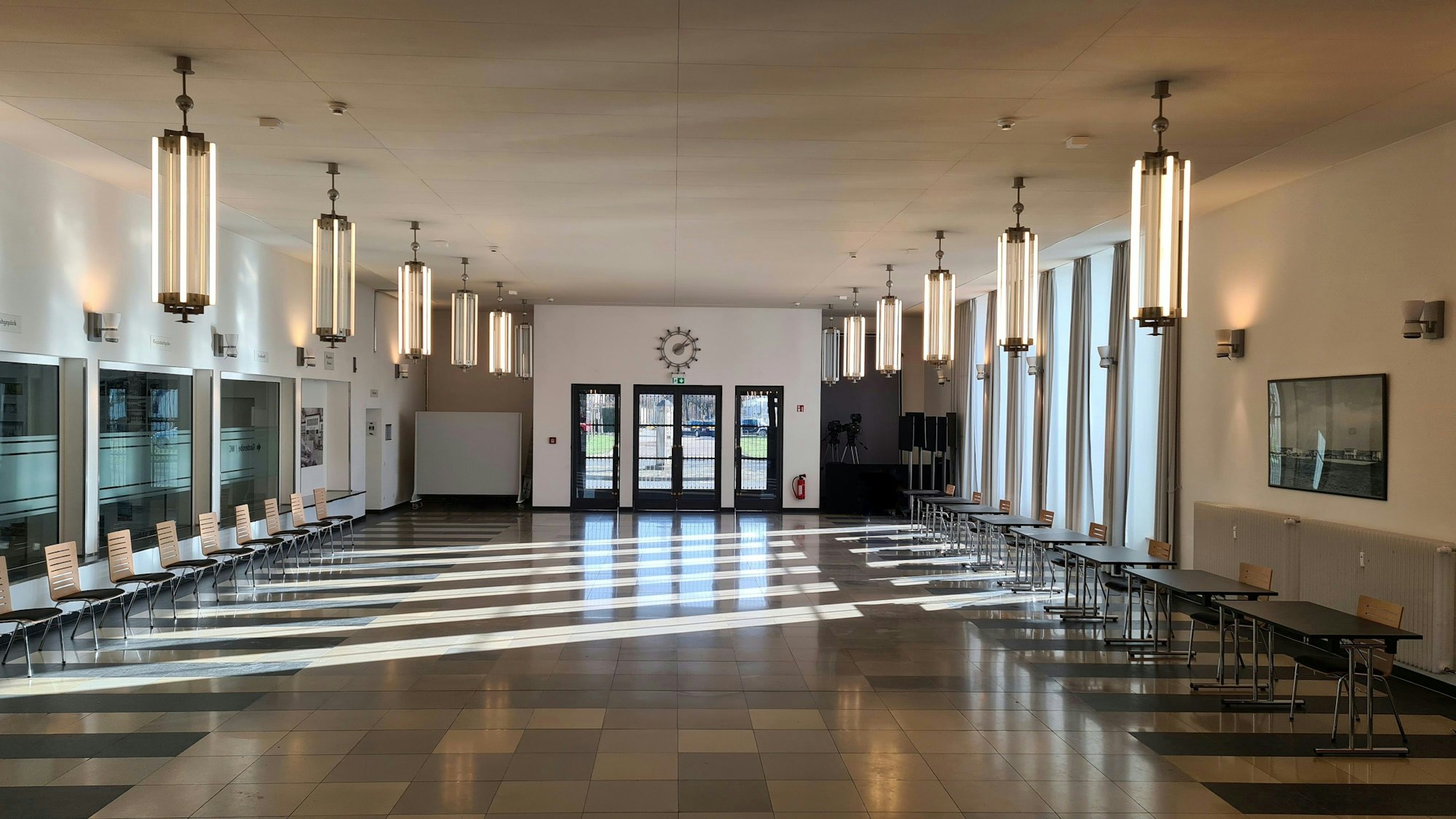 Eine lichtdurchflutete Empfangshalle eines alten Flughafens mit Stühlen an einer und Tischen an der anderen Wand ist zu sehen.