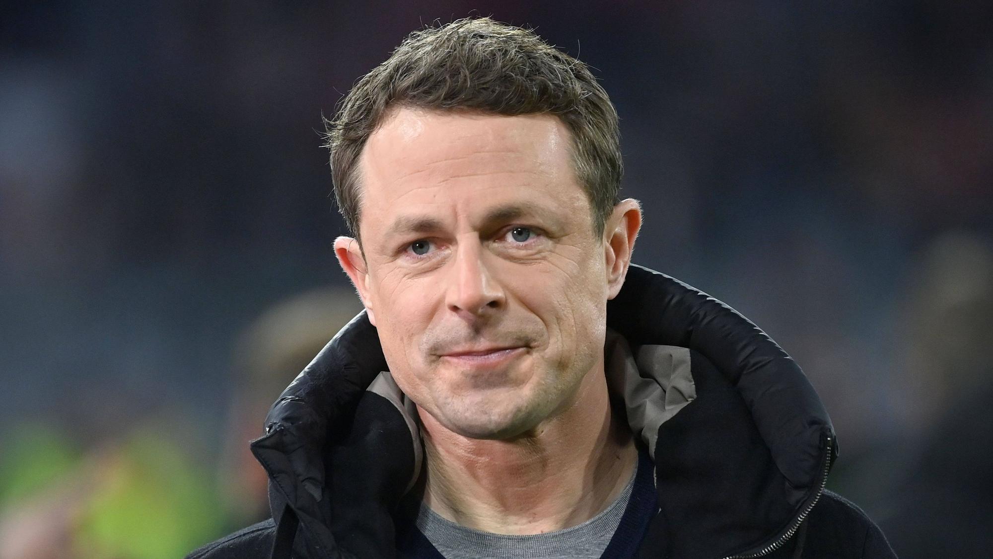 Moderator Alexander Bommes blickt bei einer ARD-Übertragung während eines Fußballspiels in die Kamera. Er trägt eine schwarze Jacke und einen grauen Pullover.