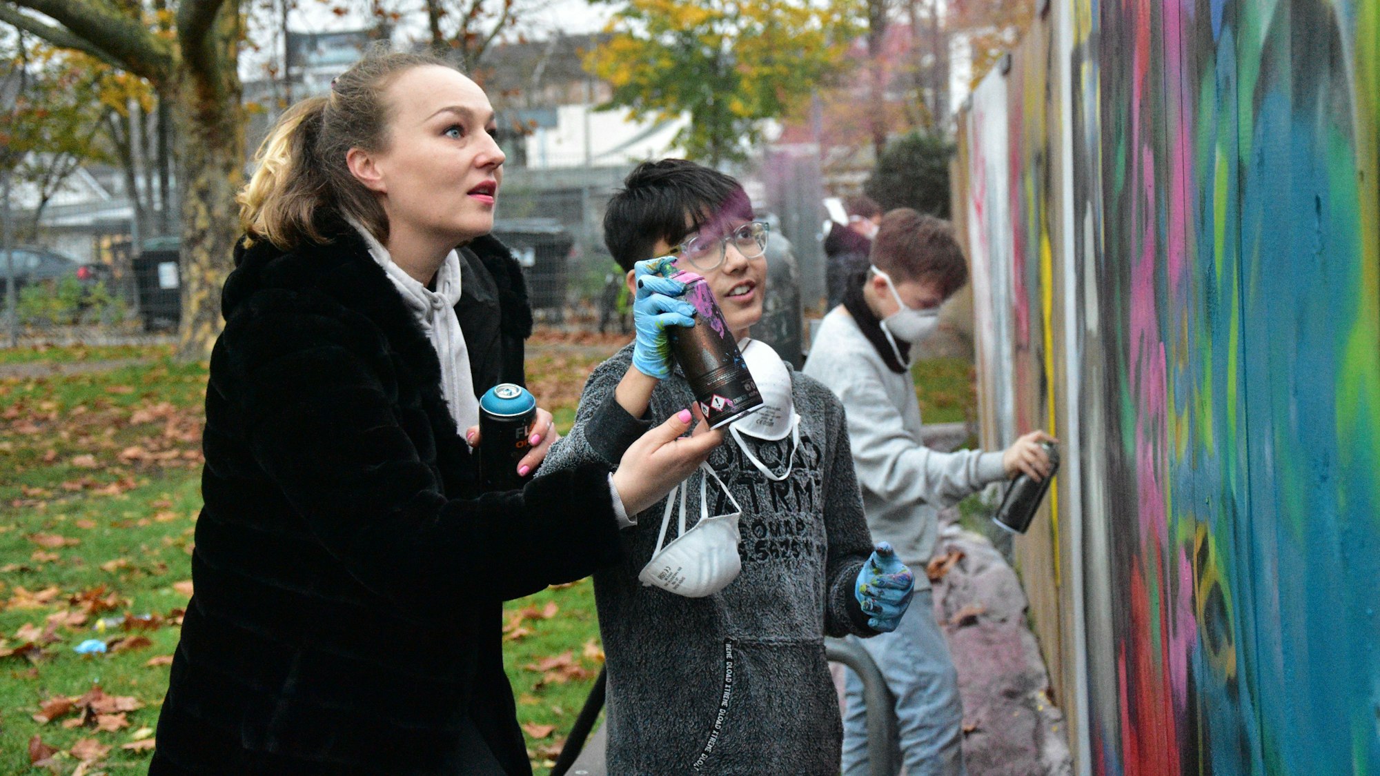 Frau und Schulkind sprühen auf bunte Graffiti-Wand.