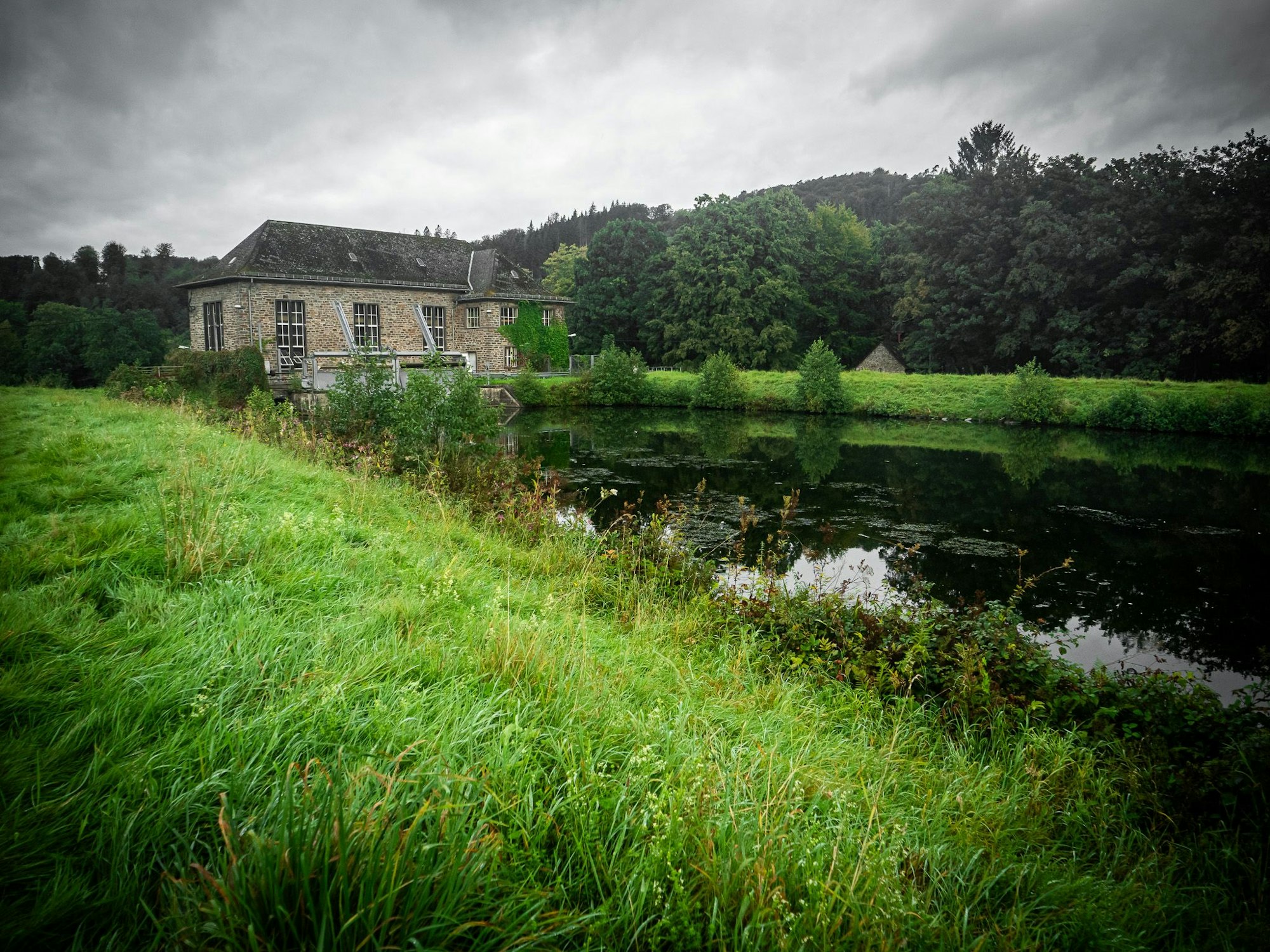Ein Bruchsteingebäude steht in grüner Landschaft an einem Stauarm der Agger in Engelskirchen.