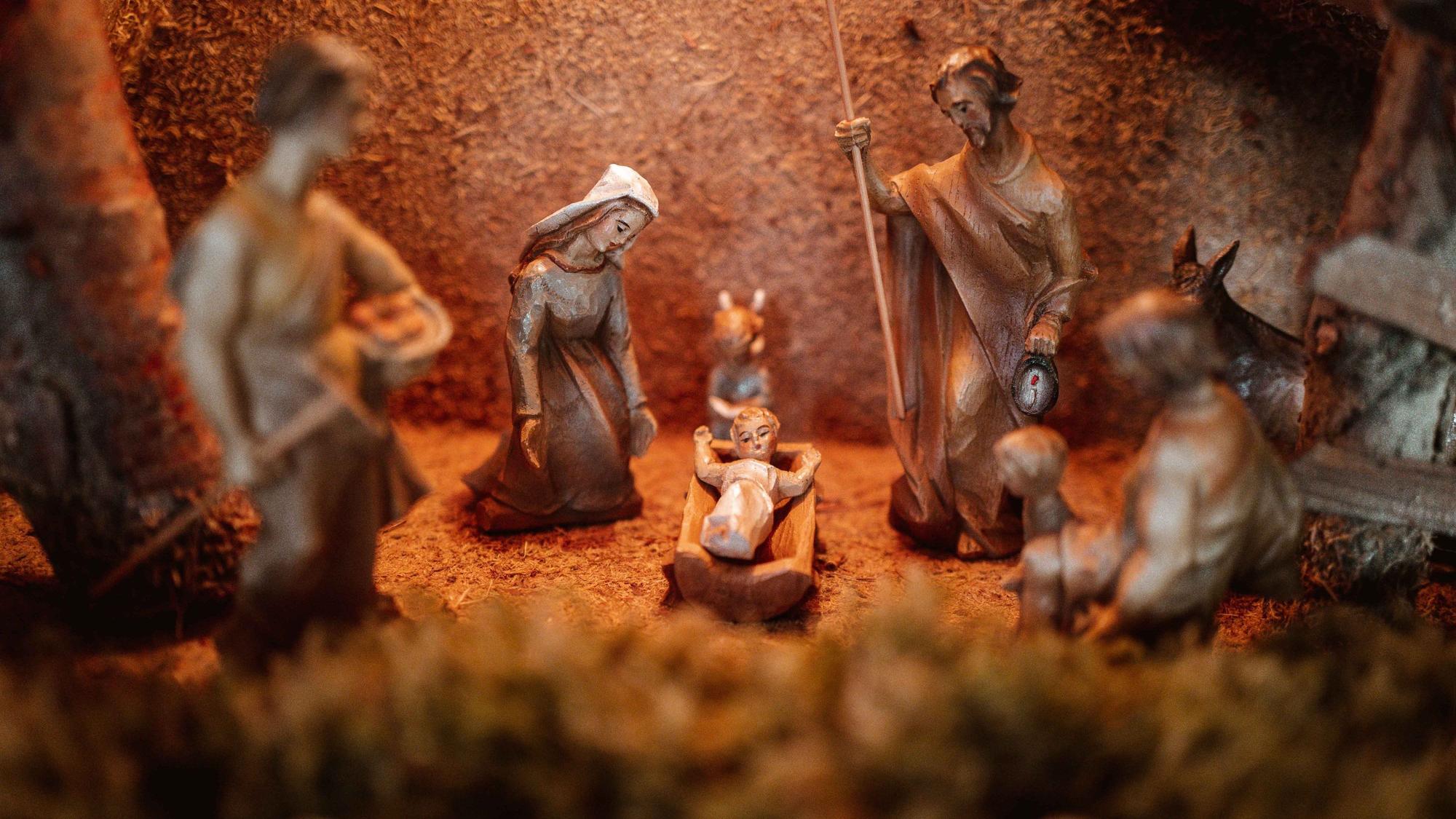 Das Bild zeigt eine Krippe. Links kniet die Figur der Maria, rechts steht Joseph mit einem Stab und in der Mitte liegt das Jesuskind in einer Krippe.