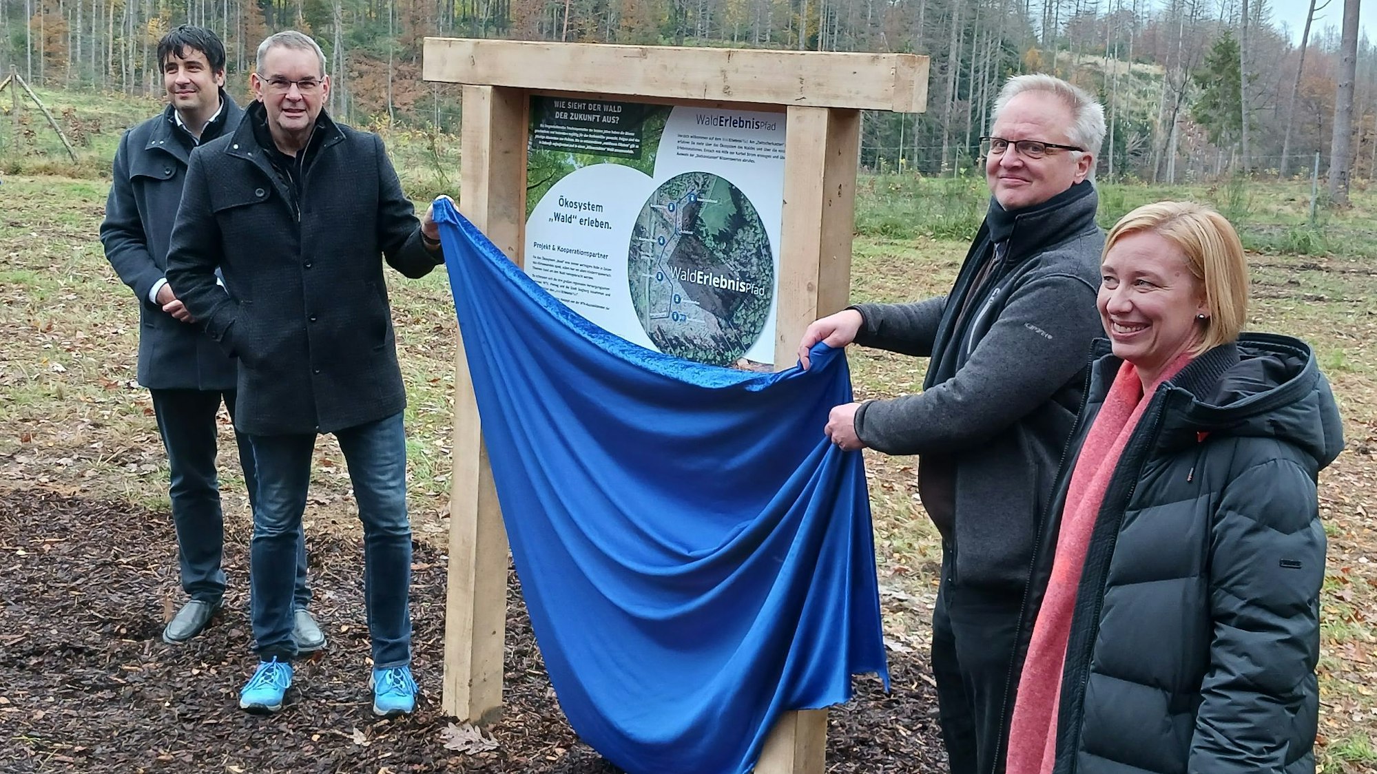 Drei Männer und eine Frau an einer Infotafel, zwei Männer enthüllen die Tafel, indem sie ein blaues Tuch wegziehen
