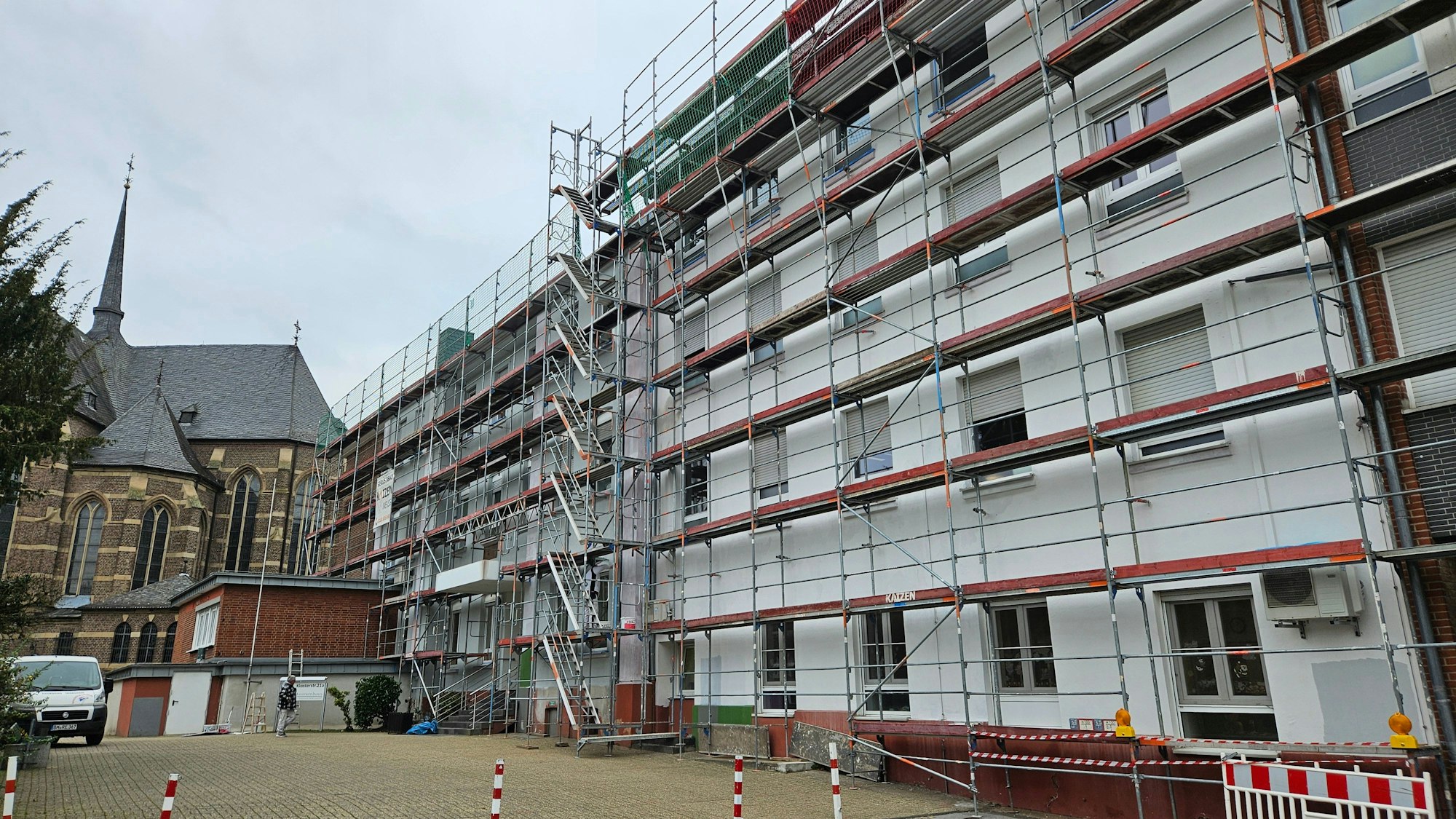 Ein großes helles Gebäude mit mehreren Stockwerken ist eingerüstet, im Hintergrund steht eine Backsteinkirche.
