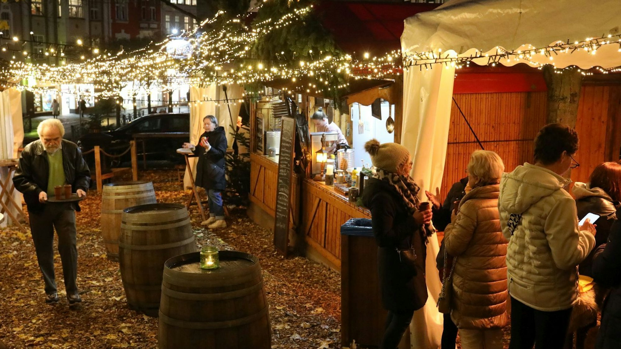 Große Holzweinfässer, die als Stehtische dienen, stehen zwischen den beiden Buden des „Kaminzimmers“. Ein Mann trägt ein Tablett mit zwei Tongläsern.