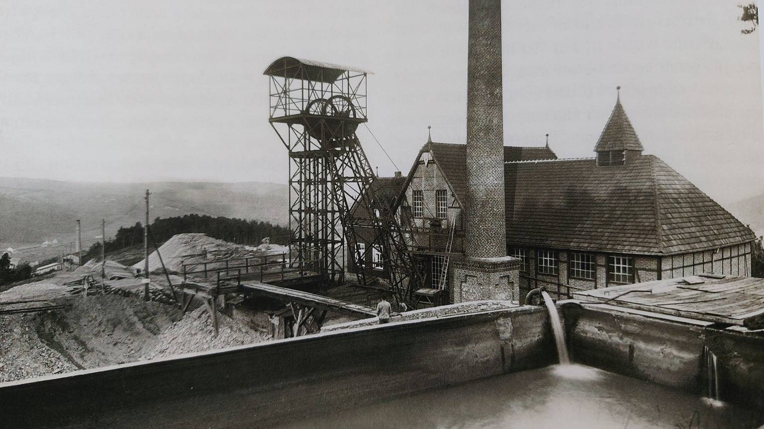 Historische Aufnahme einer alten Grubenanlage mit Förderturm und Schornstein, davor Halden.